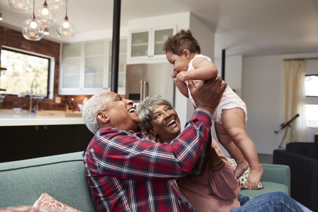 Grandparents Sitting On Sofa Playing With Baby Granddaughter At Home